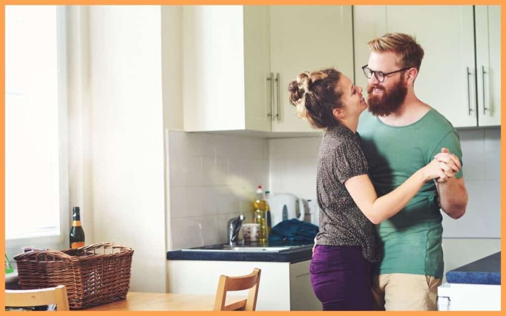 Two parents find solace in each other's arms, dancing in their kitchen amidst the chaos of parenthood. 