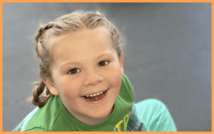 A child swinging on a swing set, their hair flying in the air, while others watch and cheer at The Village Playspace.